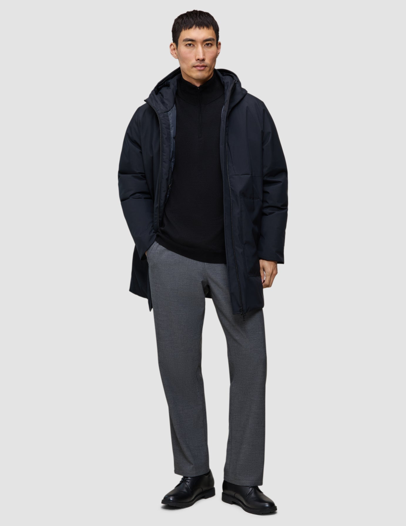 A man wearing a black Utility Jacket Black over a black zip-up sweater, grey trousers, and black shoes.